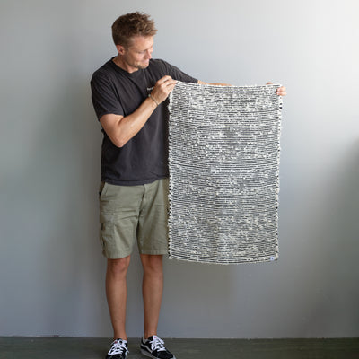 Petit tapis de 60x90cm tenu par une personne, en tissu éponge blanc avec des rayures noires, tissé à la main au Portugal.