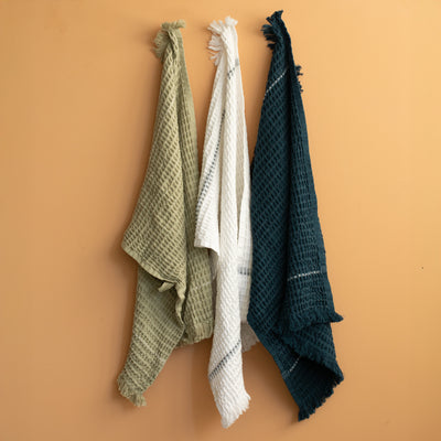 Trois serviettes gaufrées en olive, blanc et bleu marine, suspendues devant un mur de couleur terracotta