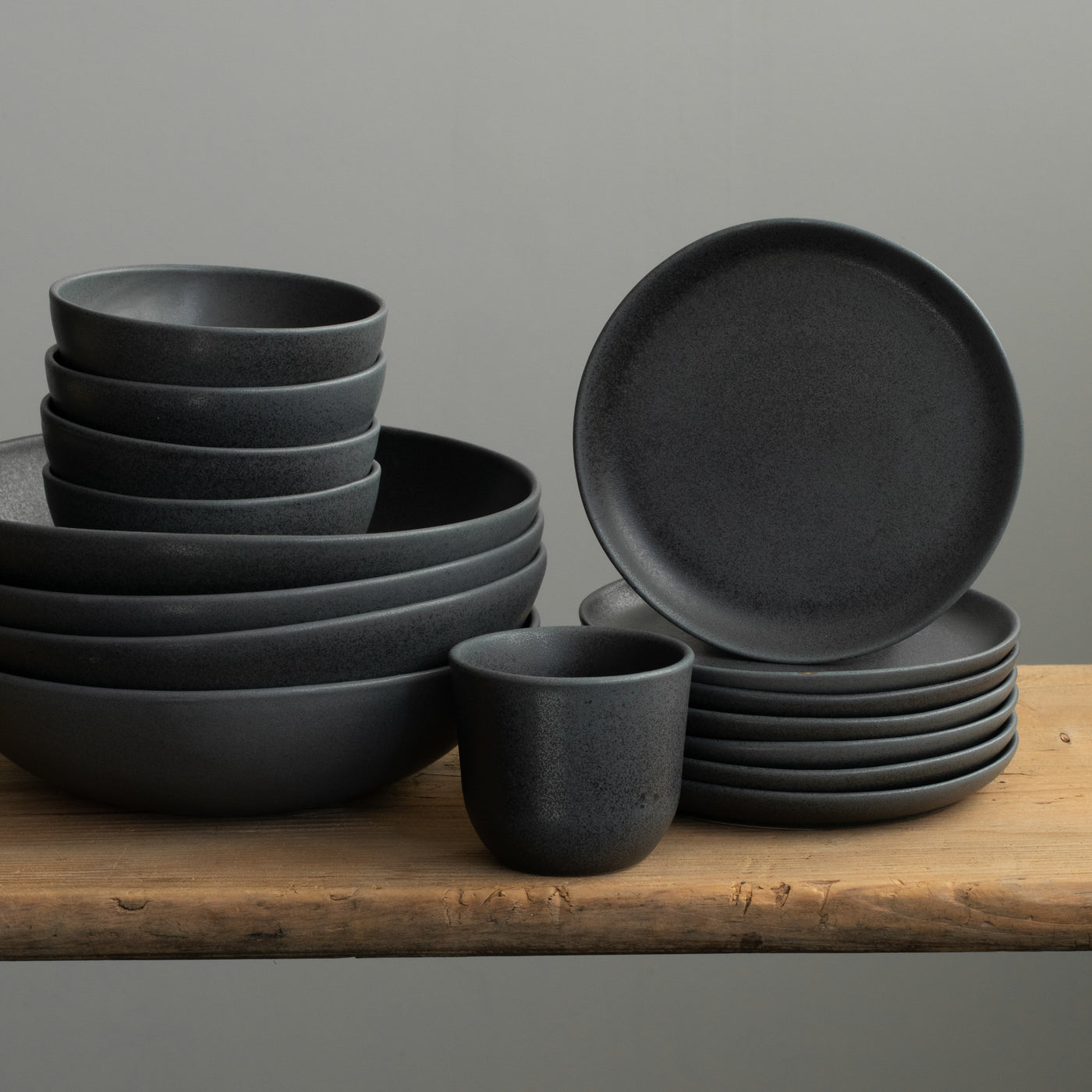 Vaisselle en grès noir mat disposée sur un plateau en bois. On y voit des assiettes à pâtes, des bols à snacks, des assiettes à pain et une tasse à cappuccino.