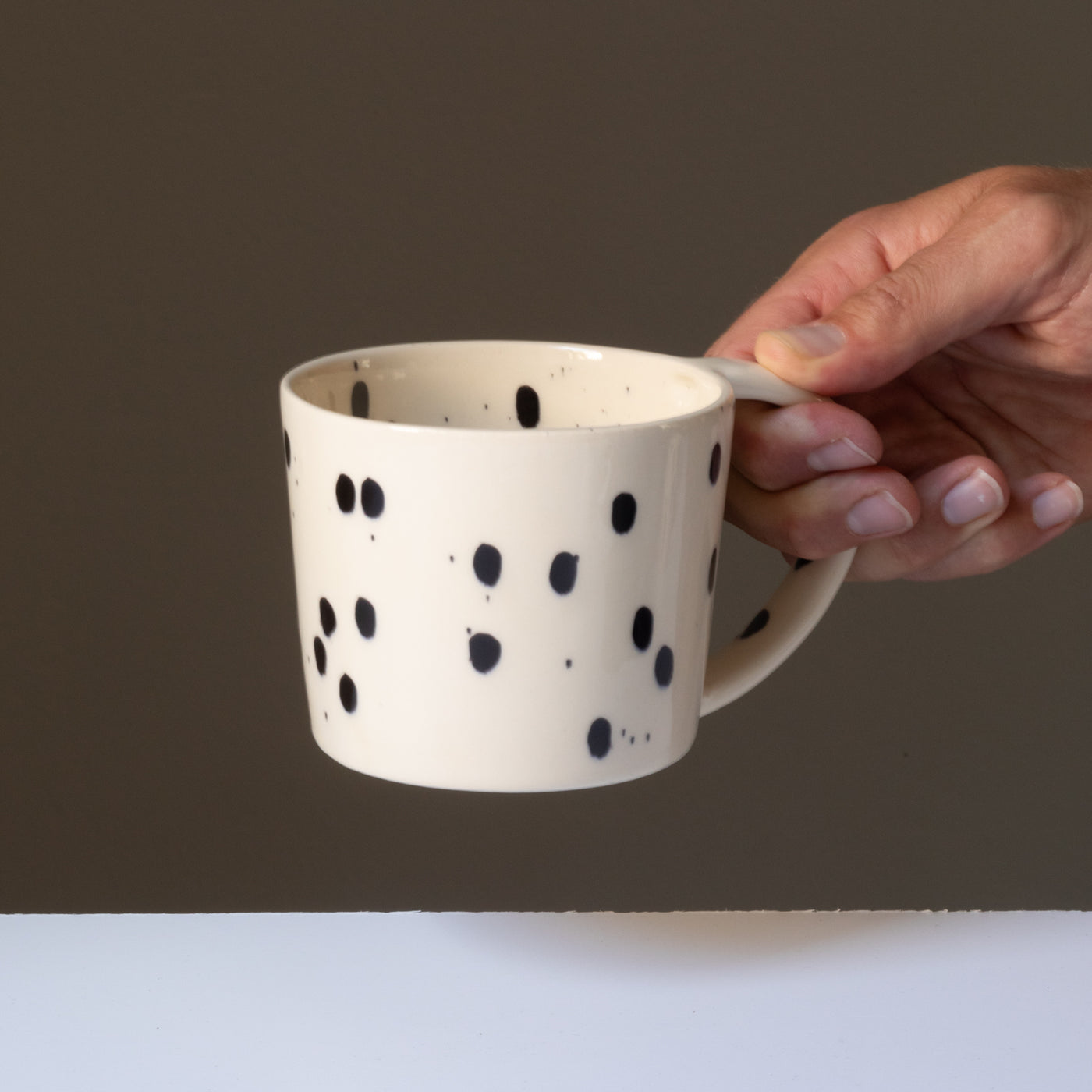 La tasse à thé de couleur crème avec des touches de noir est tenue par une main au niveau de la grande anse.