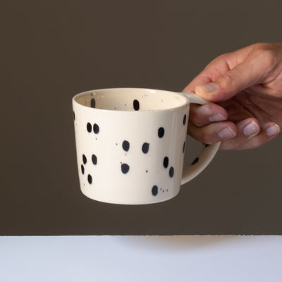 La tasse à thé de couleur crème avec des touches de noir est tenue par une main au niveau de la grande anse.