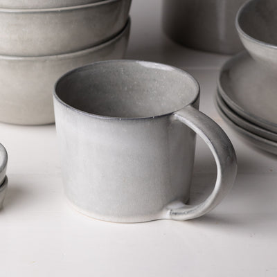 Au premier plan, une grande tasse à thé avec une anse en grès émaillé réactif gris clair. A l'arrière-plan, de grandes assiettes et des bols à muesli.